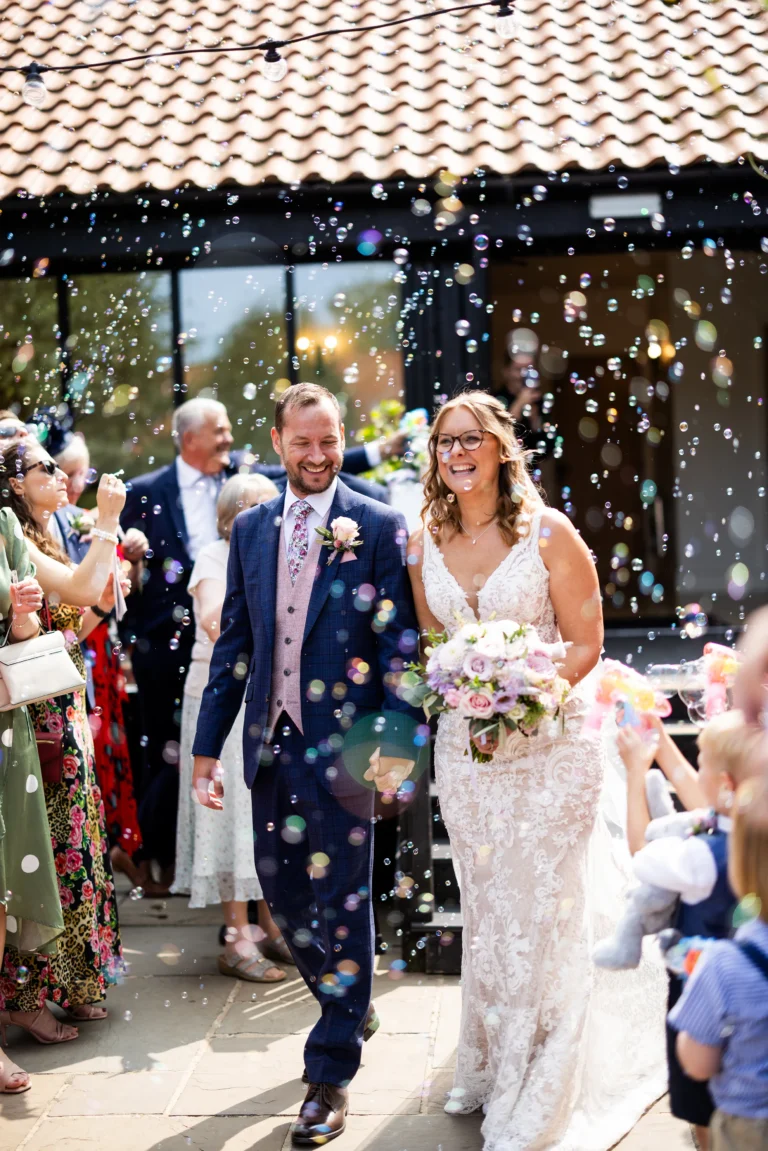 Bride and groom walking through bubble confetti, smiling and holding hands, captured by wedding photographer.